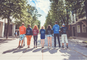 Laisvės alėjoje stovintys, į fotografą nugaras atsukę, 7 studentai su skirtingų fakultetų atstovybių džemperiais.