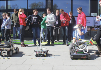 In the background we see a lot of students and in front we have three constructed robots.