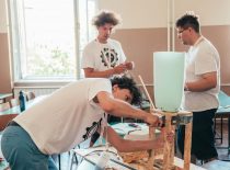 Trys studentai konstruoja meninę instaliaciją iš medienos ir plastiko plokščių.