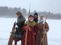Žieminiais viduramžių rūbais apsirengusios dvi moterys ir du vyrai, Viena iš moterų rankoje laiko lanką, o kita žeberklą. Vienas iš vyrų rankoje turi medinį įnagį.