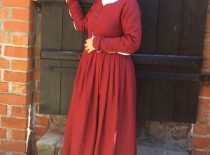 Raudona Viduramžių suknele pasipuošusi moteris stovi prie senovinių durų.
