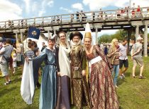 Keturios moterys apsirengusios aukšto luomo viduramžiškais apdarais šventėje. Už moterų nugaros matosi daugiau šventės dalyvių, tiltas, prekiautojų palapinės.