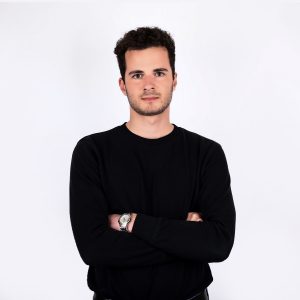 Trumpų, tamsių plaukų studentas, juodu džemperiu, sunėręs ant krūtinės rankas, ant riešo laikrodis.