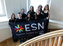 Laiptų viduryje stovi žmonių grupė, laikanti vėliavą ir &scaron;ypsosi. Už jų matyti du langai.