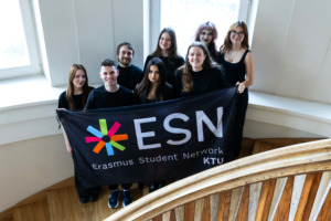 Laiptų viduryje stovi žmonių grupė, laikanti vėliavą ir &scaron;ypsosi. Už jų matyti du langai.