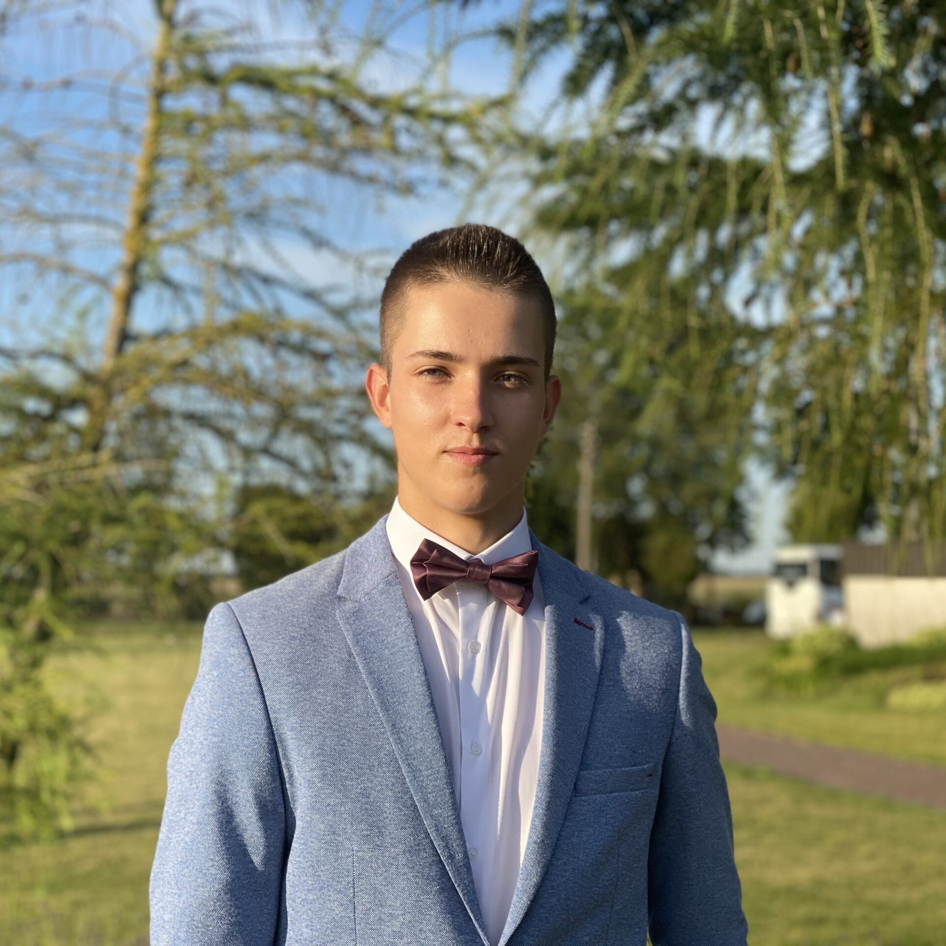 Studentas, trunpais, juodais plaukais, dėvintis marškinius, pilką švarką ir varlyte miško fone.