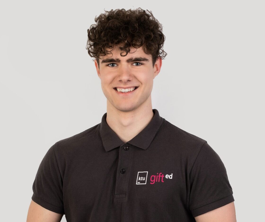 Vaikinas tamsiai rudais plaukais bei tamsiai pilkai marškinėliais stovi baltame fone.