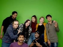 Aštuoni studentai pozuojantys fotografui prie žalios sienos.