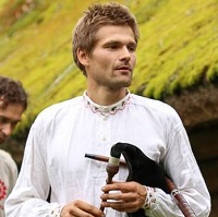 Šviesių, trumpų plaukų vyras, baltais lininiais marškiniais, rankose laikantis kelis muzikos instrumentus.