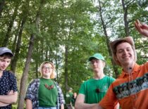 „Jaunystės“ kolektyvo nariai Valčių žygyje. 4 choristai šypsosi ir pozuoja. Fone matoma žaluma ir medžiai.