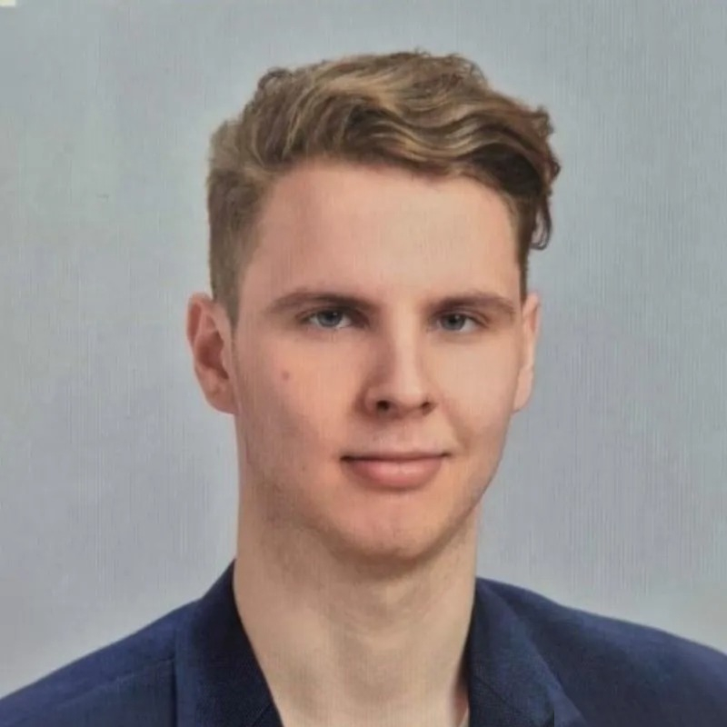 Portretinė nuotrauka jauno vaikino su trumpais šviesiais plaukais ir šoniniu sklastymu. Jis vilki tamsų švarką ir pozuoja prieš šviesų foną su lengva šypsena.