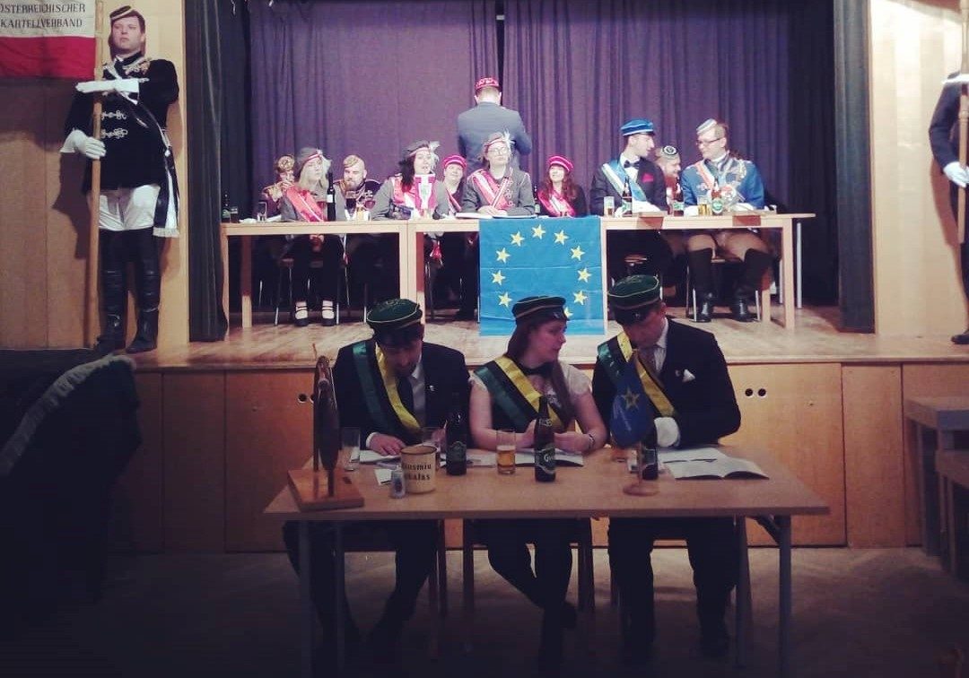 Salėje ant scenos prie ilgo stalo sėdi būrys studentų, stalo priekyje Europos sąjungos vėliava, kairėje pusėje studentas laikantis plakatą, nulipus nuo scenos prie stalo sėdi trys studnetai ir šnekasi tarpusavyje.