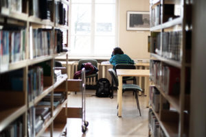 KTU biblioteka. Iš kairės ir dešinės pilnos knygų lentynos. Centre, žaliu džemperiu vilkinti studentė sėdinti prie stalo.