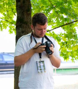 Vaikinas su baltais marškinėliais žaliame medžio fone, nuleidęs galvą žiūri į rankose esantį fotoaparatą,