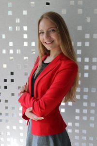 Plačiai besišypsanti, ties liemeniu sukryžiuotomis rankomis šviesių plaukų mergina vilkinti raudoną švarkelį ir pilką sijoną.
