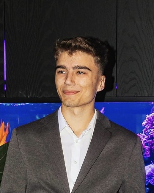 Jaunas vaikinas su trumpais rudais plaukais vilki elegantišką pilką kostiumą ir baltus marškinius. Jis šypsosi ir žvelgia į šoną. Fone matomas akvariumas su ryškiaspalvėmis žuvimis ir koralais, taip pat juodos spintelės su violetiniu apšvietimu.