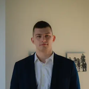 Laimingai atrodantis vyras trumpais plaukais, vilkintis baltus marškinius, mėlyną švarką, šviesus fonas su kabančiais paveikslais.