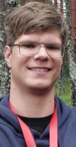 Jaunas vaikinas, su akiniais, apsirengęs pilką džemperį ir ant kaklo raudoną pakapubą.